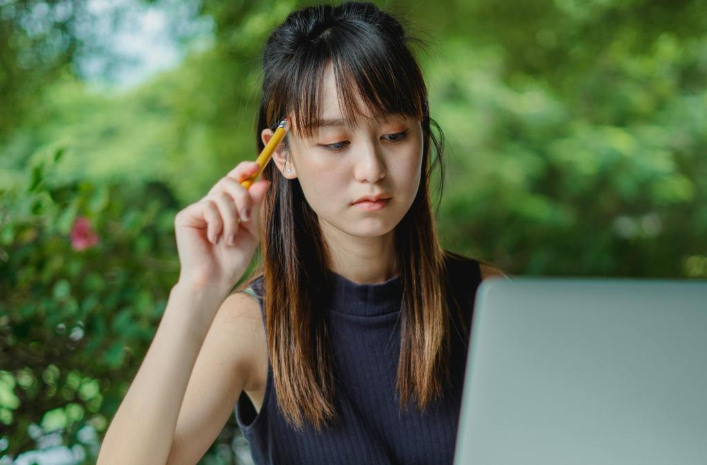 Young Asian lady holding a pen to her head while looking at a laptop